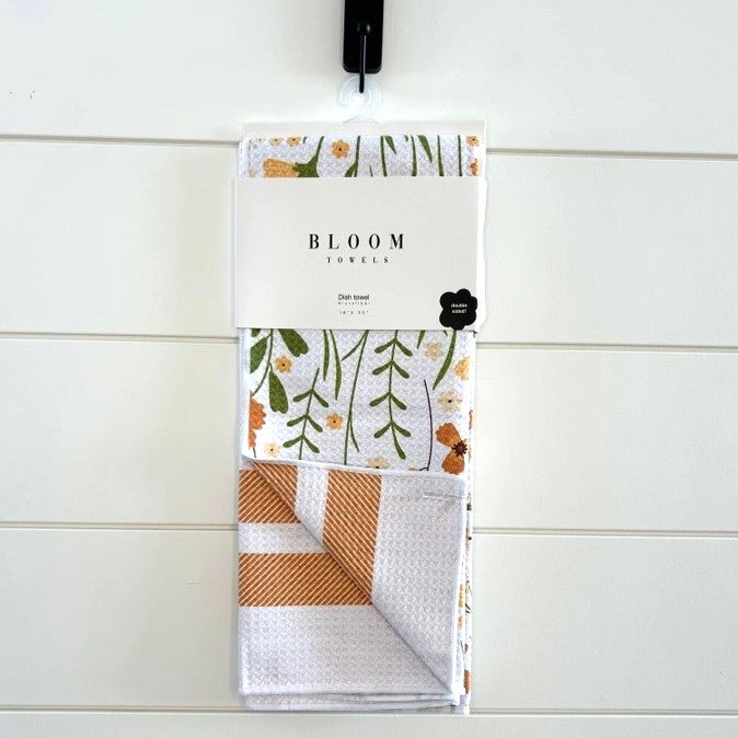 Set of two folded towels with floral and checkered patterns in packaging labeled 'Bloom Towels' on a white wooden background.