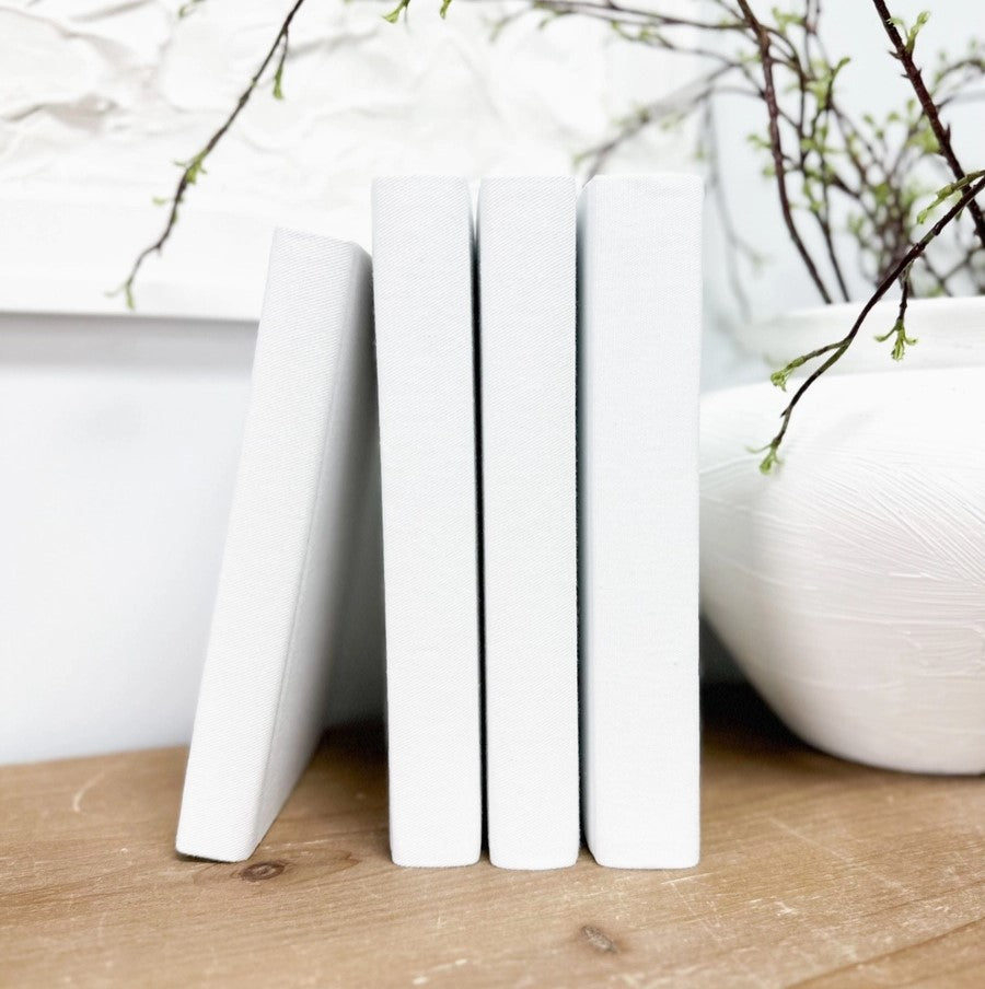 A white linen book with a smooth spine, displayed vertically on a wooden surface with a blurred background featuring branches and a bowl.