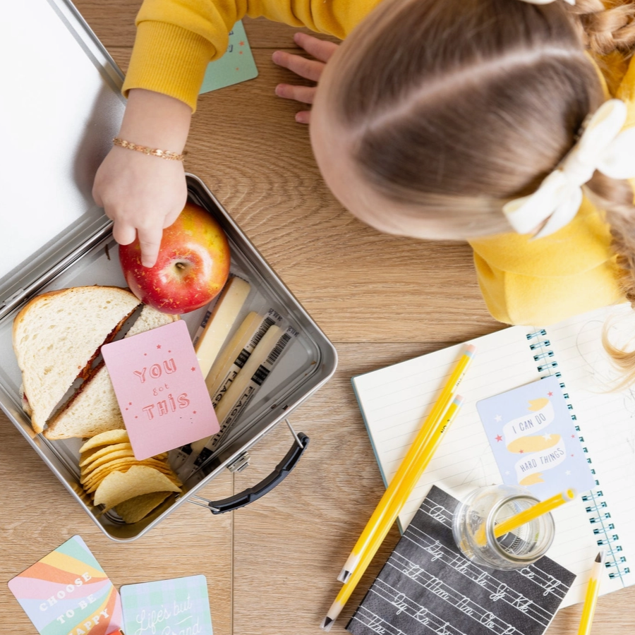 Child reaching into a lunchbox with an apple, sandwich, and snacks on a wooden table.