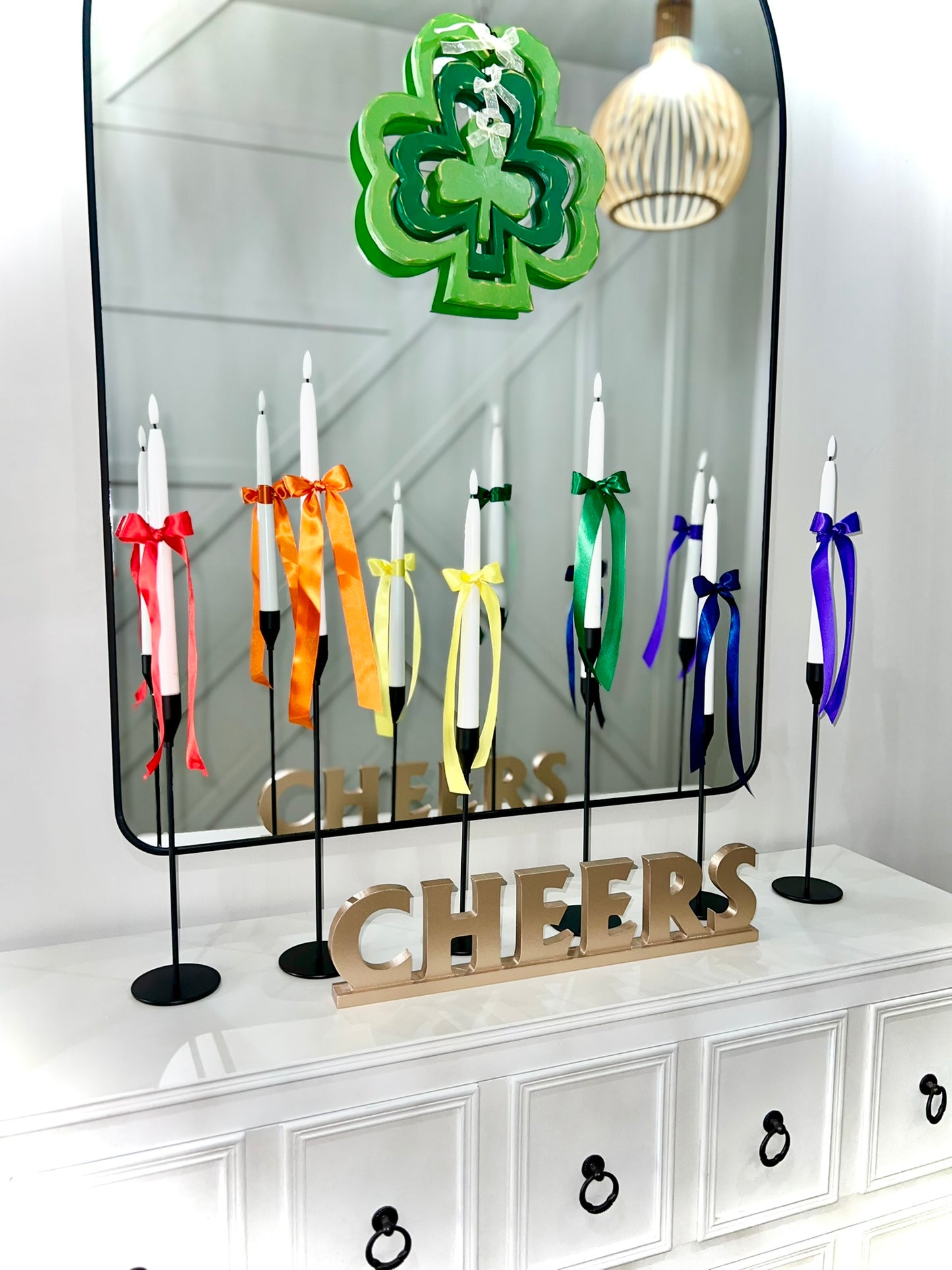 A set of colorful satin ribbons tied to candlesticks, with each ribbon a different color corresponding to the colors of the rainbow, displayed in front of a mirror with a 'CHEERS' sign.