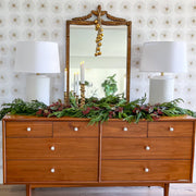 Wooden dresser with decorative mirror and greenery against a patterned wall.