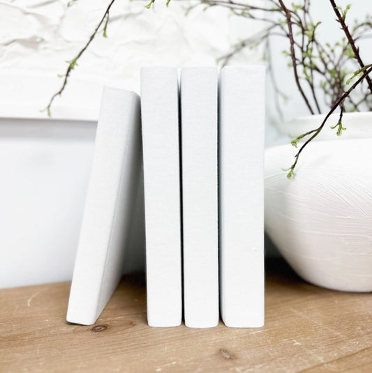A white linen book with a smooth spine, displayed vertically on a wooden surface with a blurred background featuring branches and a bowl.
