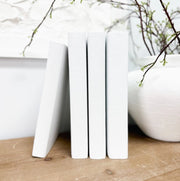 A white linen book with a smooth spine, displayed vertically on a wooden surface with a blurred background featuring branches and a bowl.