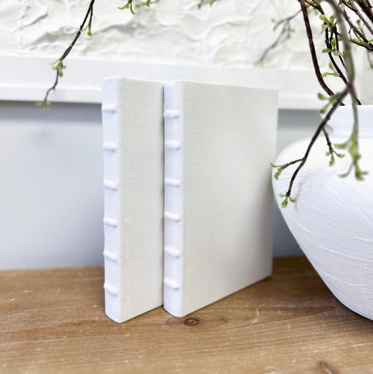 A white linen book with a wrapped spine placed upright on a wooden surface, with a decorative item partially visible in the top right corner.