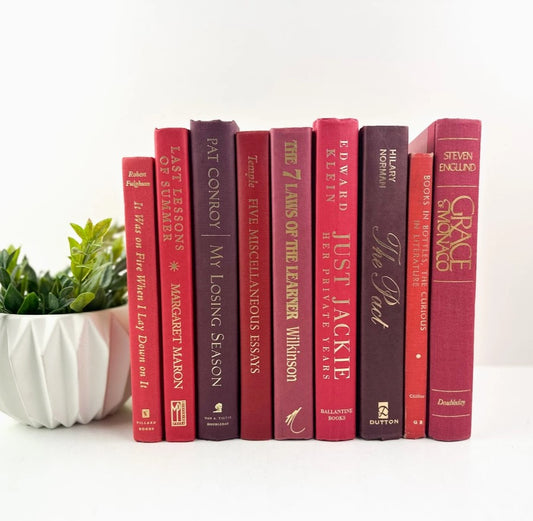 A series of decorative books arranged on a shelf with a white plant pot to the left, all with red spines and titled with variations of 'The,' ranging in size.