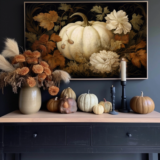 Decorative setup with pumpkins, flowers, and candles on a table against a wall with a pumpkin-themed painting.