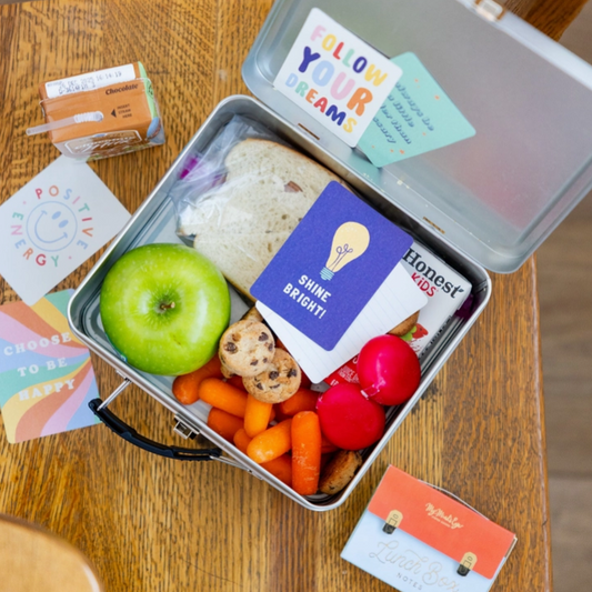Metal lunchbox with snacks and a card inside, surrounded by additional cards on a wooden surface.