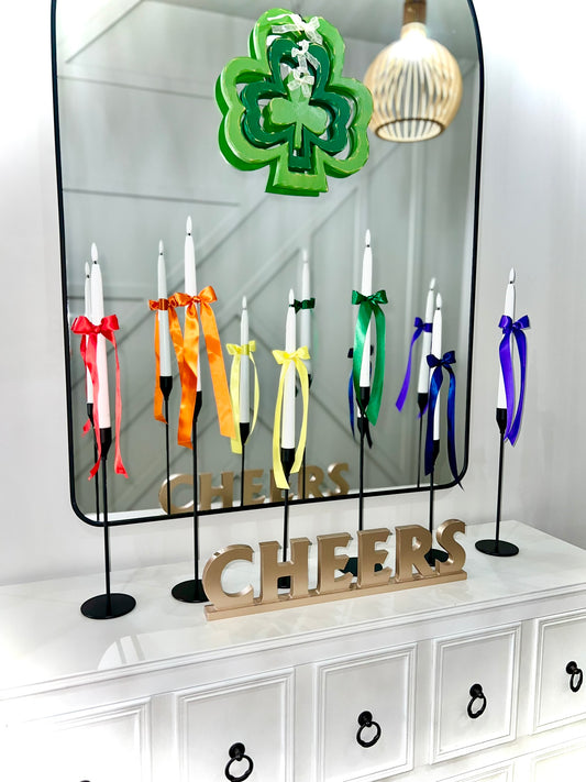 A set of colorful satin ribbons tied to candlesticks, with each ribbon a different color corresponding to the colors of the rainbow, displayed in front of a mirror with a 'CHEERS' sign.